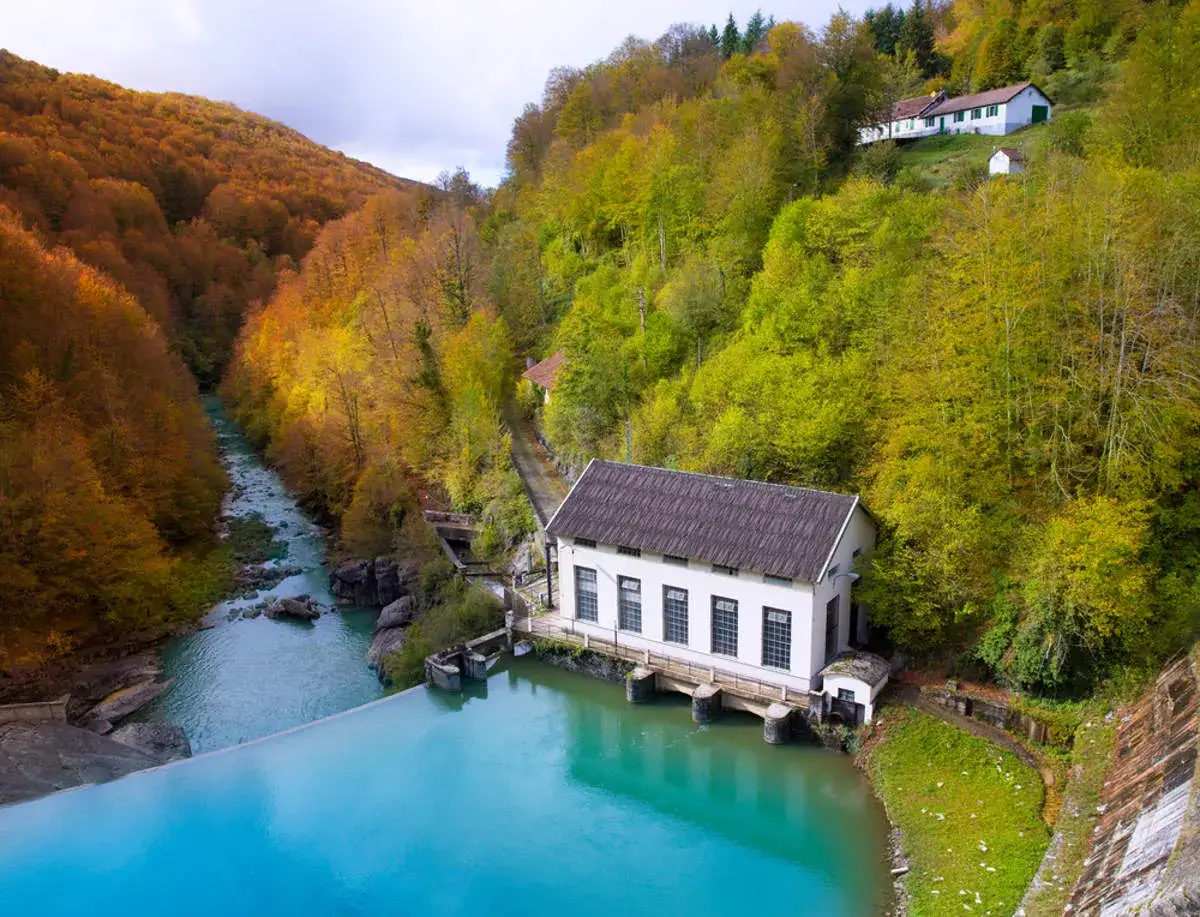 Un día de otoño en la selva de Irati, con niños jugando cerca de un río turquesa y una casa blanca entre árboles de colores vibrantes.