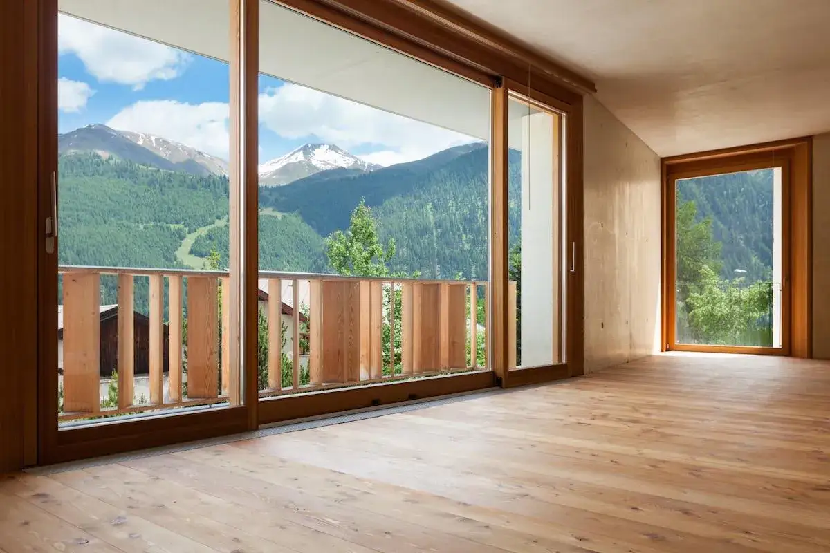 Interno con pavimento in legno e ampio arredamento con infissi marroni che incorniciano un panorama montano mozzafiato.