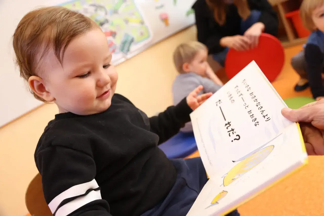 Un ni&ntilde;o peque&ntilde;o sonr&iacute;e mientras le leen un libro ilustrado, practicando ejercicios de lengua.