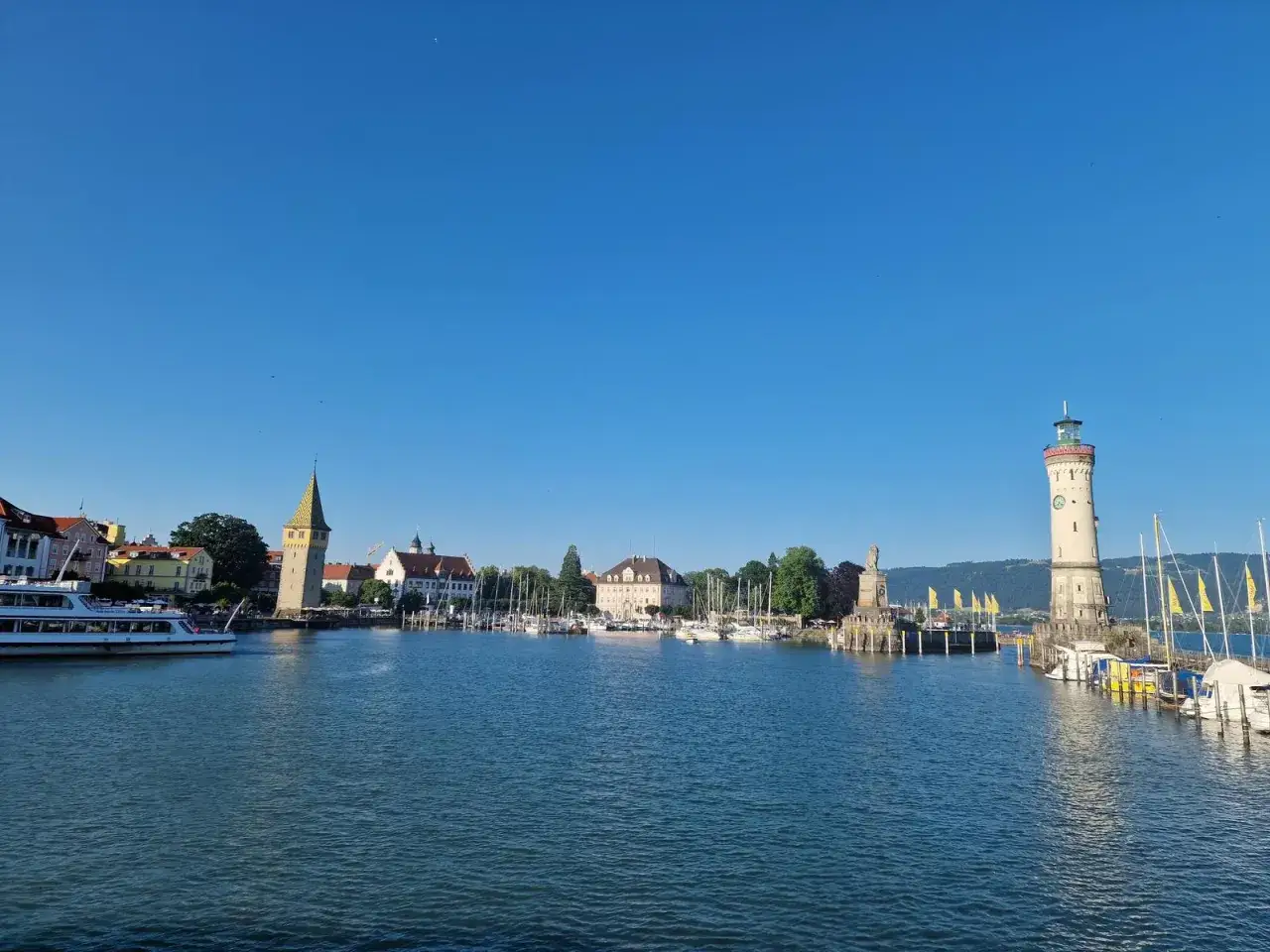 Porto con faro, torre e barche. Un assaggio della Deutsche Alpenstrasse, con il lago che riflette il cielo azzurro.