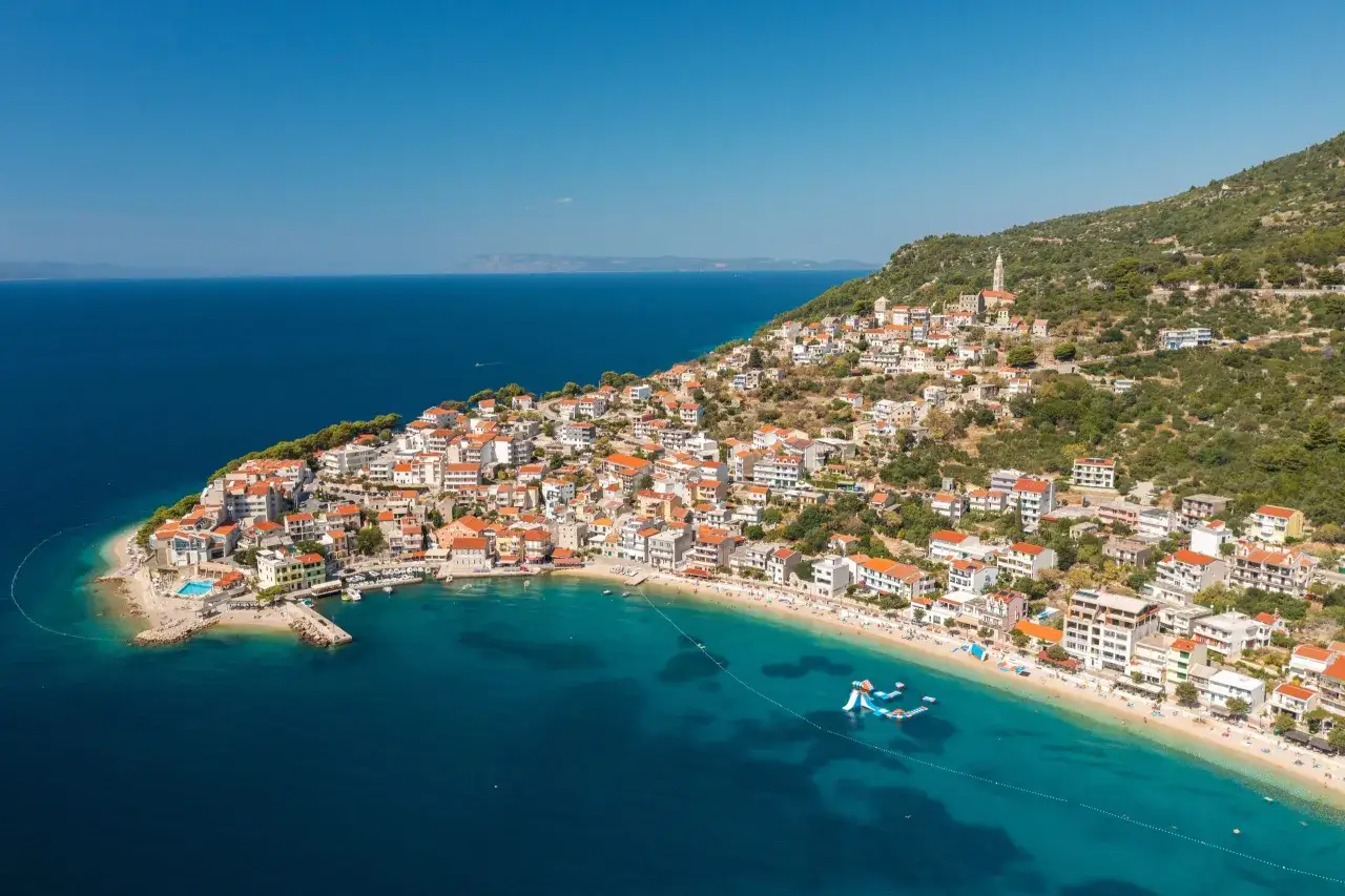 A picturesque village clings to the hillside along the Balkan coast, with terracotta roofs cascading down to a turquoise bay dotted with boats and swimmers.