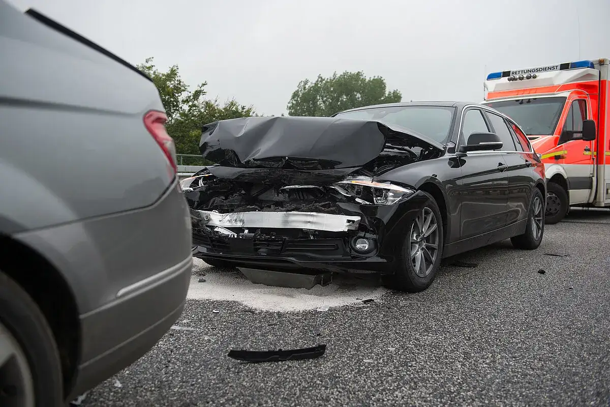 Ein schwarzer PKW hat einen schweren Frontschaden, die Motorhaube ist stark verformt. Ein Rettungswagen steht daneben.
