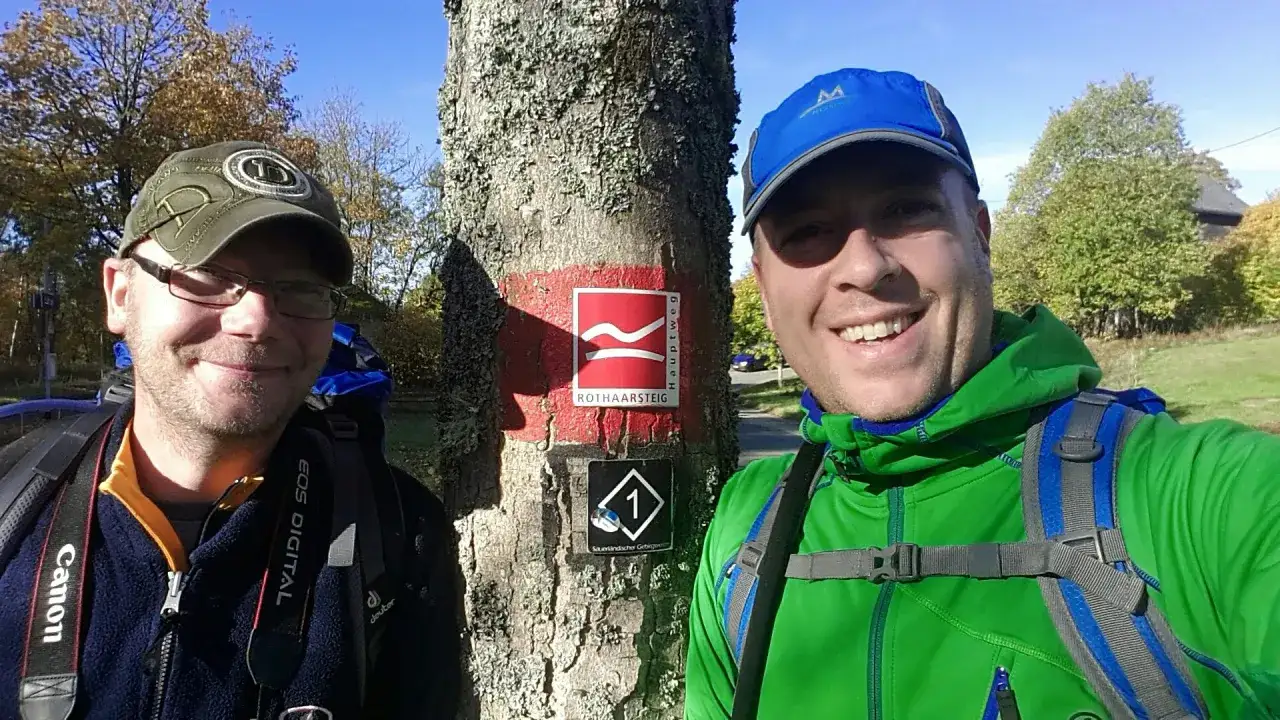 Zwei Wanderer l&auml;cheln in die Kamera, neben ihnen ein Baum mit dem Zeichen des Rothaarsteigs. Komoot-Nutzer auf Tour.