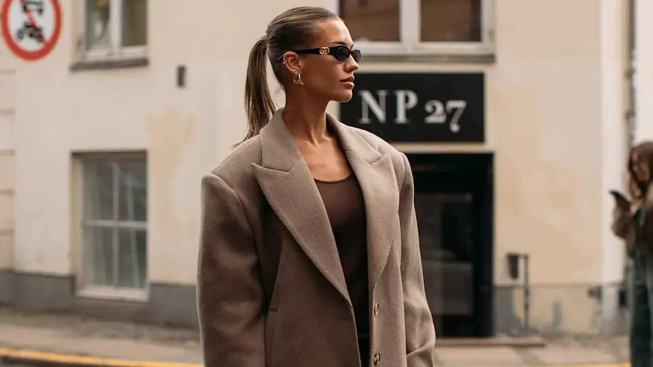 Mujer con gafas de sol y un abrigo oversize, su mirada es demure mientras posa en la calle.