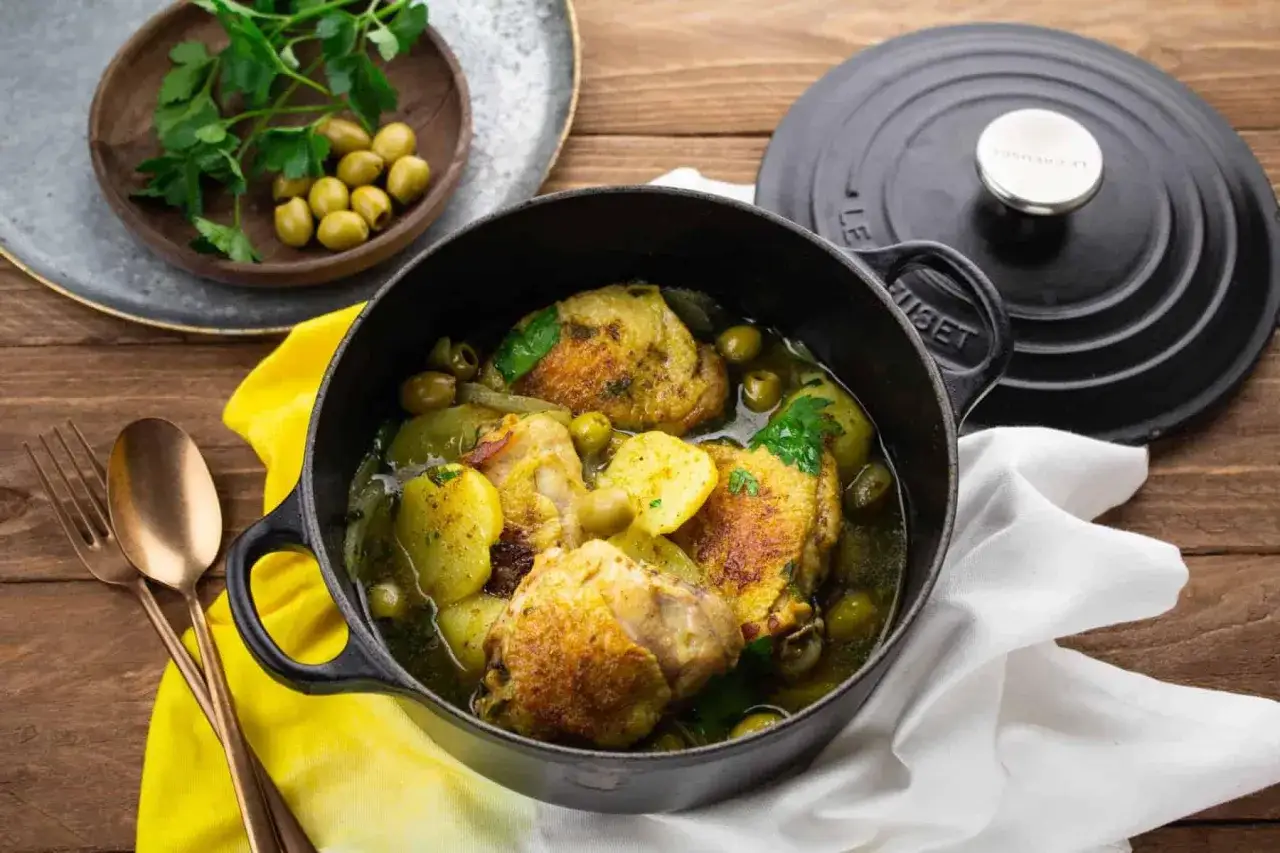 Un plat de poulet aux olives mijoté dans une cocotte en fonte, garni de pommes de terre et de persil frais.