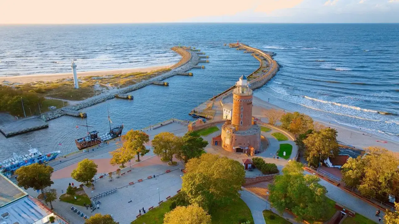 Widok na latarnię morską Kołobrzeg, port z żaglowcem i nowoczesną jednostką, plażę i morze.