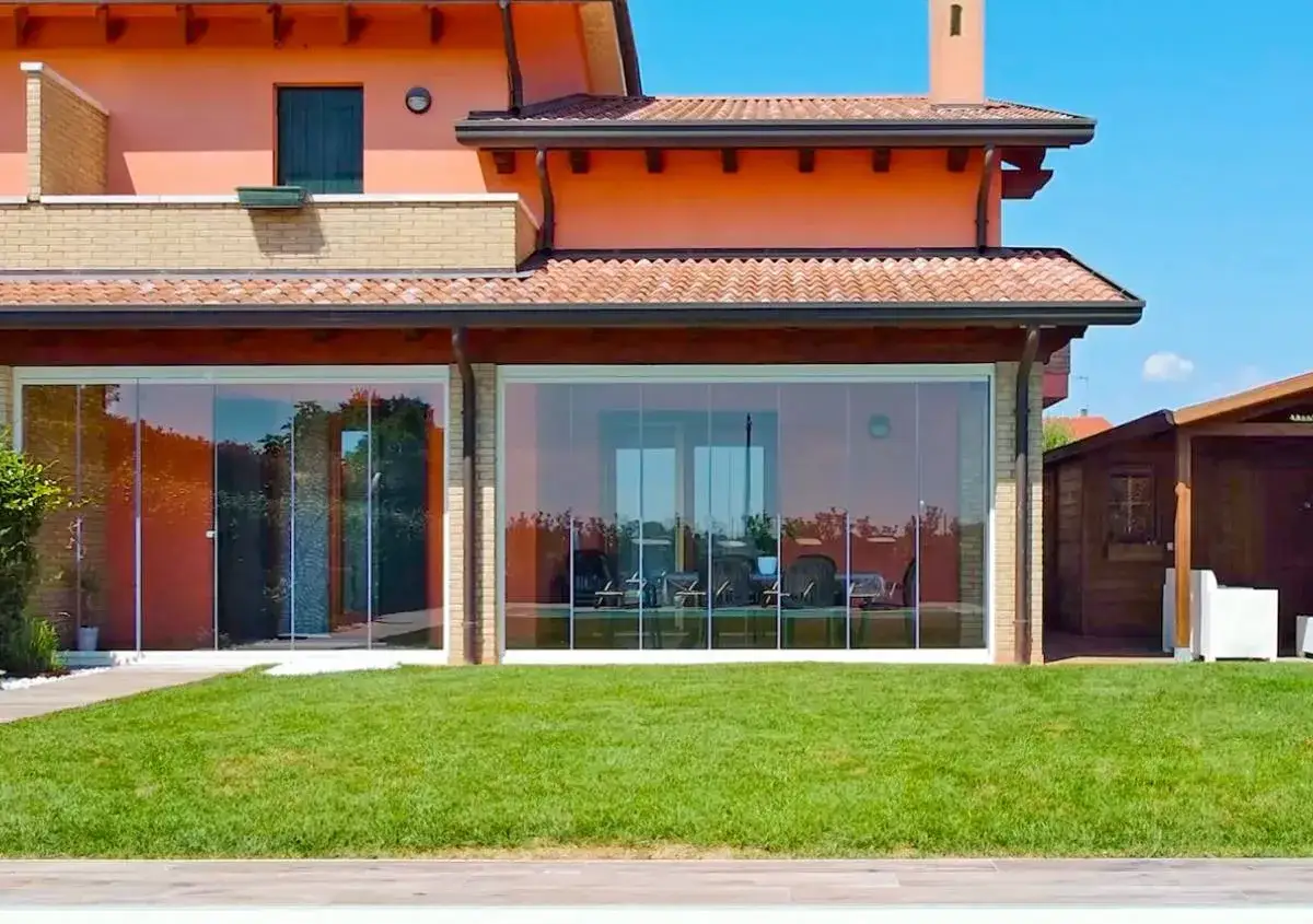 Moderna casa con ampie vetrate che creano eleganti architetture in vetro, riflettendo il verde del giardino e il cielo azzurro.