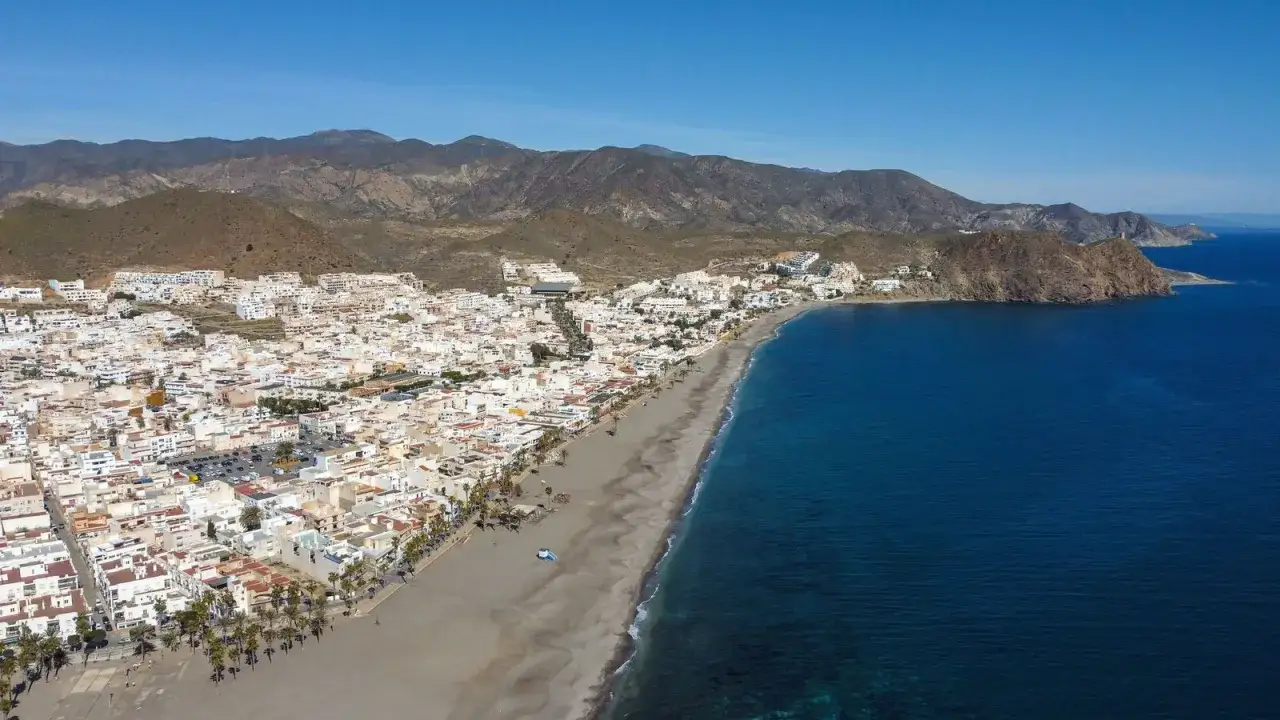 Vista aérea de la **playa de las Salinicas**, con casas blancas y montañas al fondo. El mar azul contrasta con la arena oscura.