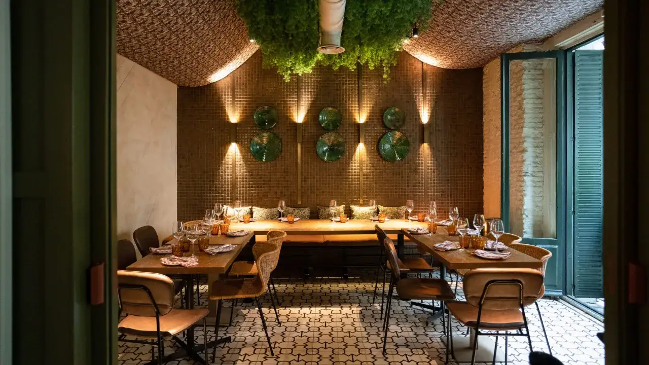 Un elegante ambiente restaurante con mesas puestas, sillas de cuero y mimbre, y una exuberante vegetaci&oacute;n colgante.