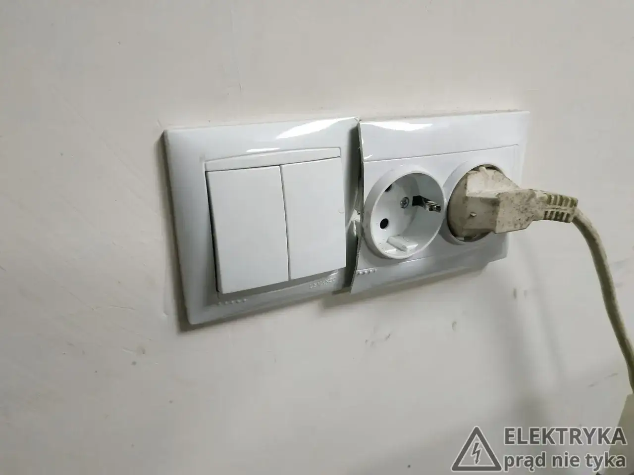 Biały montaż: włącznik światła i gniazdko elektryczne z wtyczką. Ściana w tle jest jasna, z widocznymi śladami malowania.