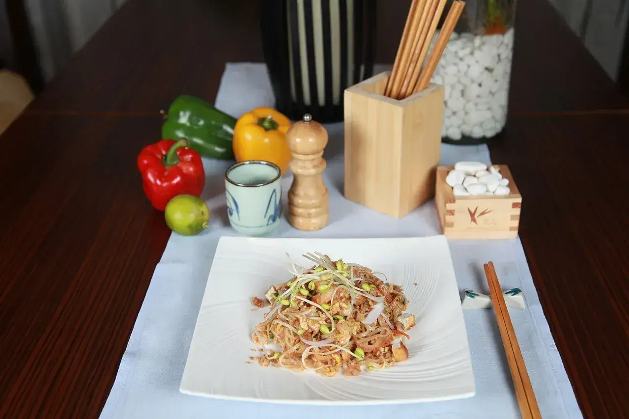 Talerz z daniem przypominającym pad thai, ozdobiony kiełkami. Obok papryki, limonka, filiżanka, młynek do pieprzu i pałeczki. Idealne jako banchan.