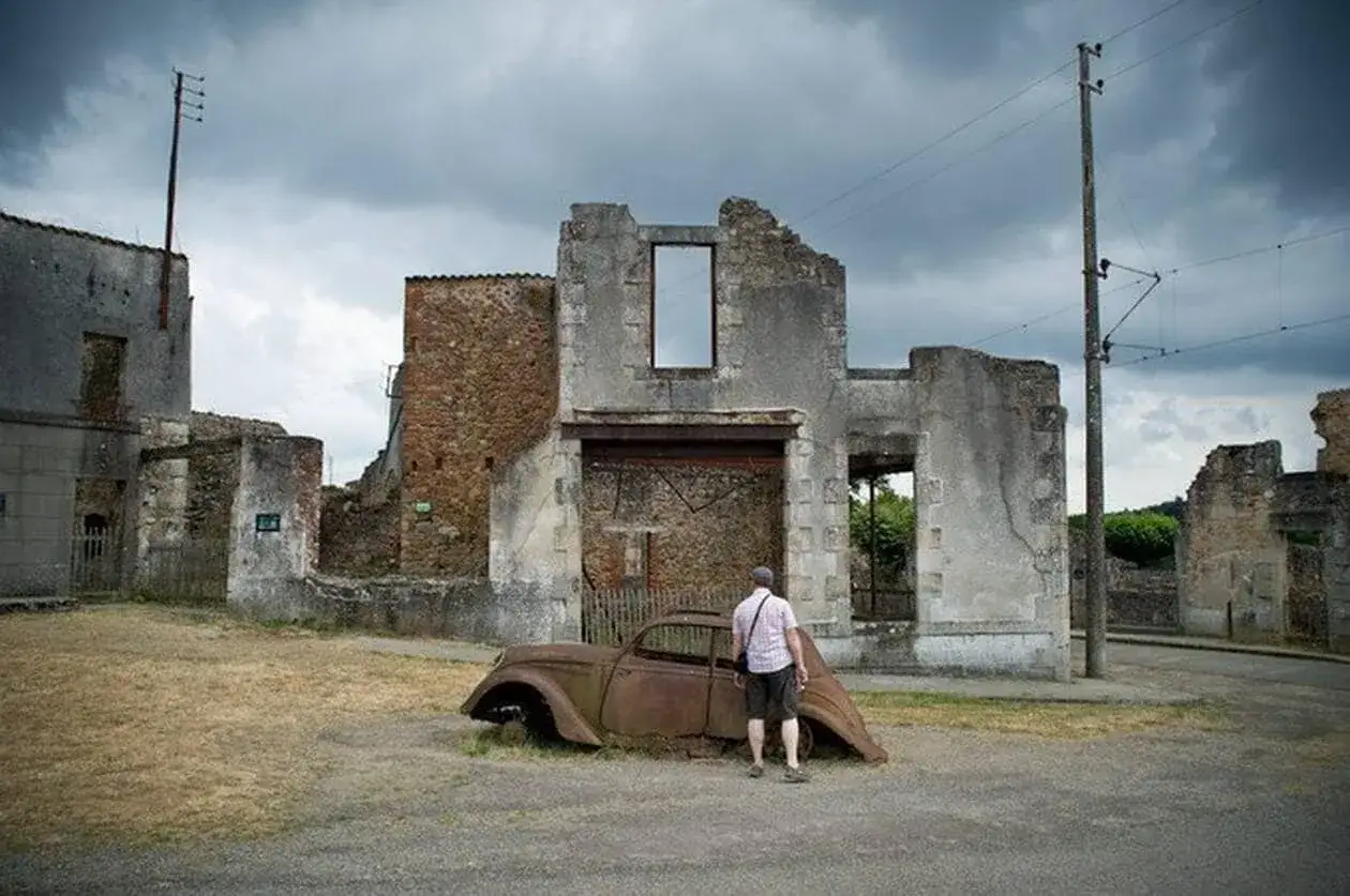 Un homme observe une vieille voiture rouill&eacute;e au milieu d'un village abandonn&eacute;, entour&eacute; de ruines sous un ciel mena&ccedil;ant.