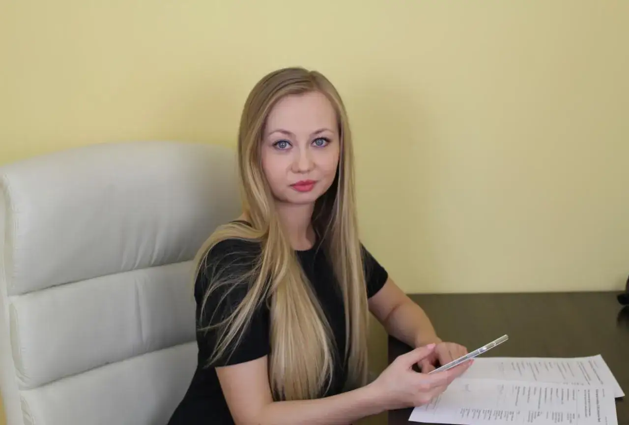 Kobieta z długimi blond włosami i niebieskimi oczami siedzi przy biurku, trzymając telefon.