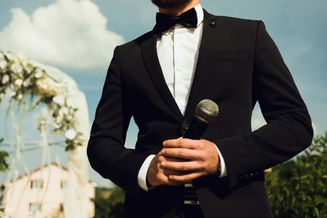 Un maestro de ceremonias con traje negro y pajarita sostiene un micrófono, listo para dar inicio a una boda al aire libre.