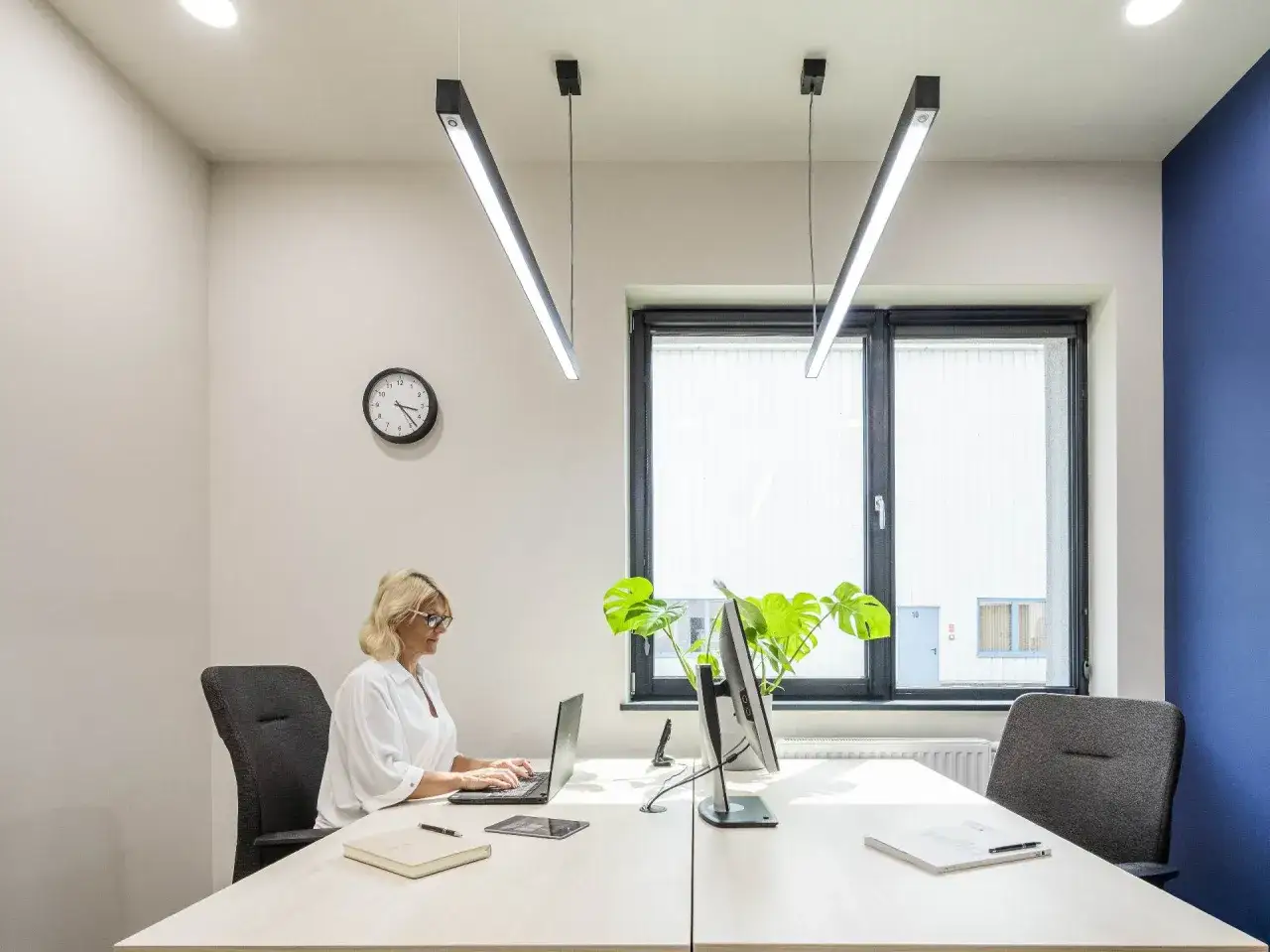 Bureau moderne avec femme travaillant sur ordinateur portable. La lumi&egrave;re naturelle et les suspensions LED cr&eacute;ent une ambiance propice au respect de la norme &eacute;clairage bureau.