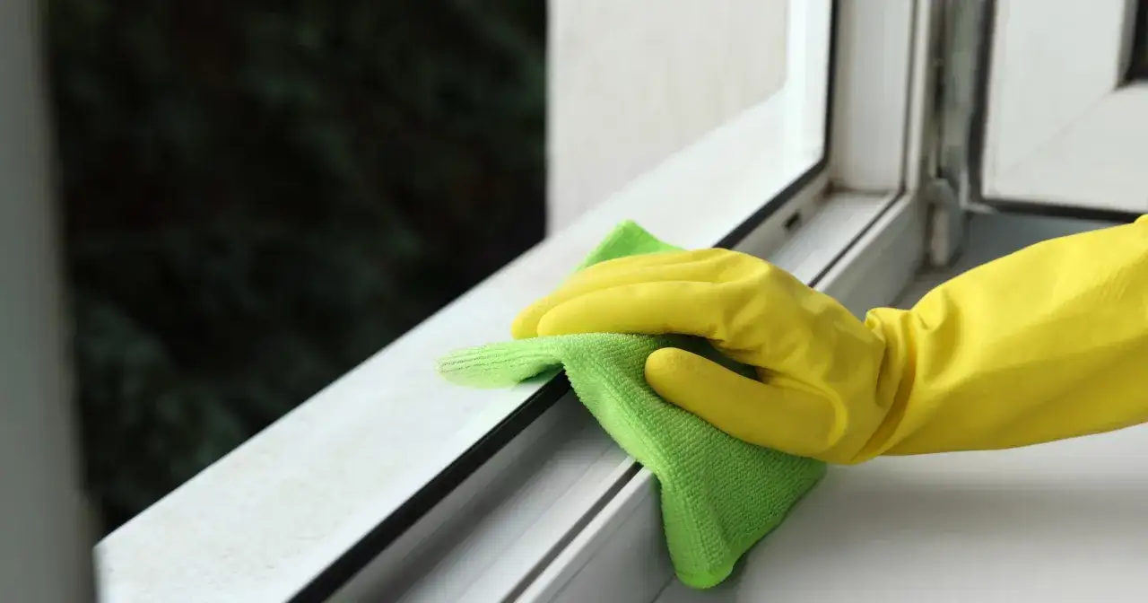 Ręka w ż&oacute;łtej rękawiczce czyści białą ramę okna zieloną ściereczką. Dowiedz się, czym wybielić okna plastikowe, by zn&oacute;w lśniły.