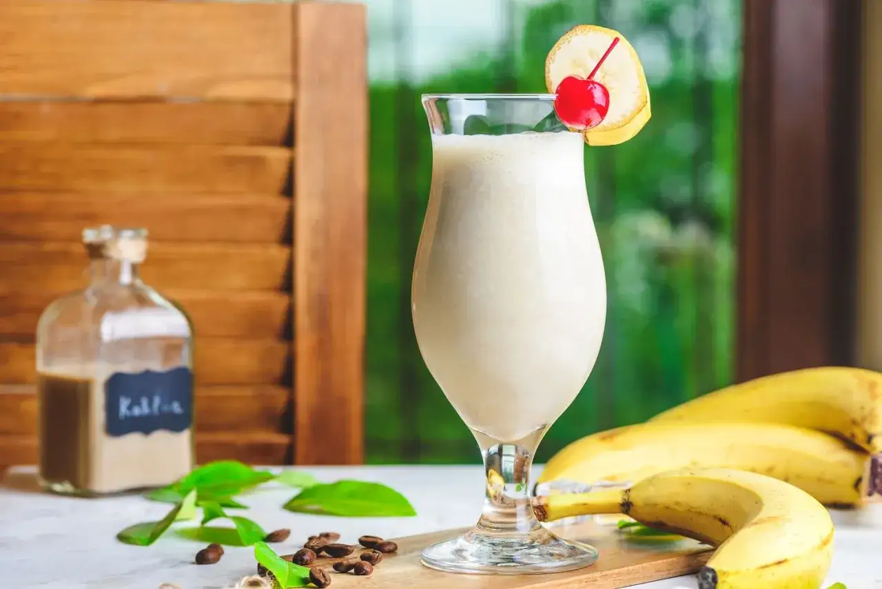 Orzeźwiający drink z sokiem bananowym, ozdobiony wiśnią i plasterkiem banana. Obok buteleczka z likierem kawowym.