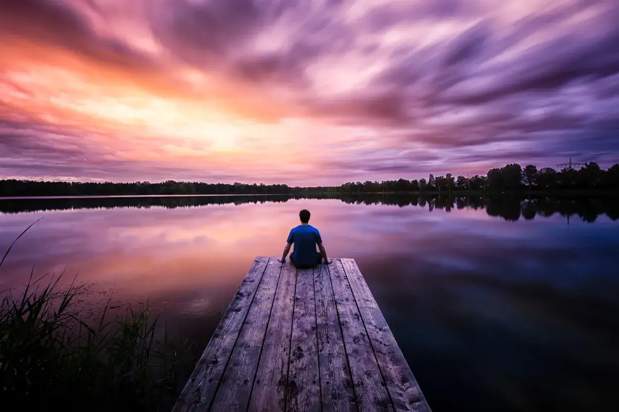 Zachód słońca nad jeziorem, człowiek siedzi na pomoście. Fotograf krajobrazu uchwycił magiczne chwile.