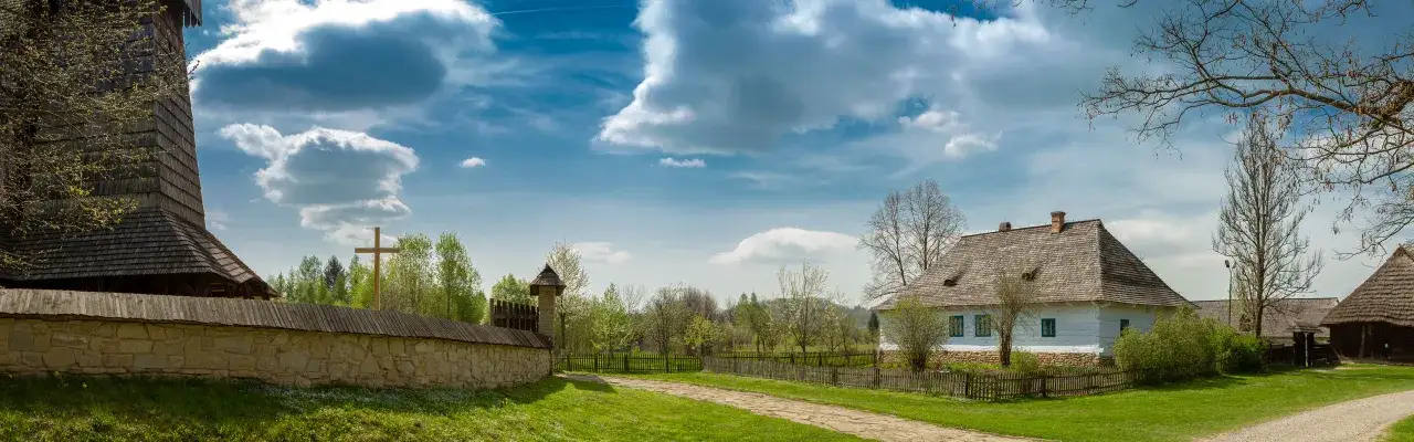 Drewniana wieża i biały dom z niebieskimi okiennicami na tle błękitnego nieba z chmurami. To największy skansen w Polsce, gdzie można poczuć ducha przeszłości.