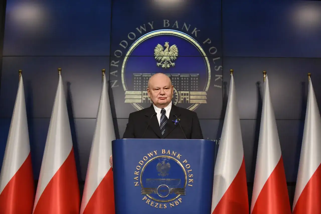 Prezes NBP Adam Glapiński przemawia, omawiając narzędzia polityki pieniężnej. Za nim godło Polski i flagi.