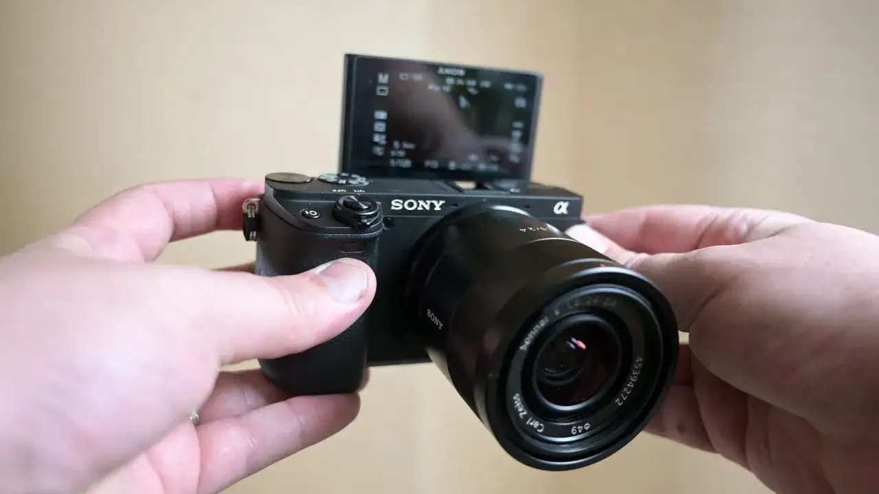 Ręce trzymają aparat Sony a6400 z obiektywem Carl Zeiss. Ekranik jest uchylony, widać menu.