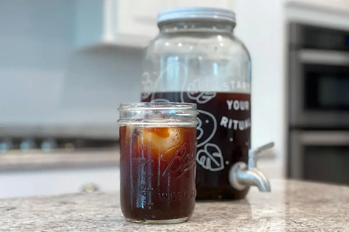 Ett glas iskaffe med is framför en stor behållare med cold brew. Perfekt för ett enkelt cold brew recept.