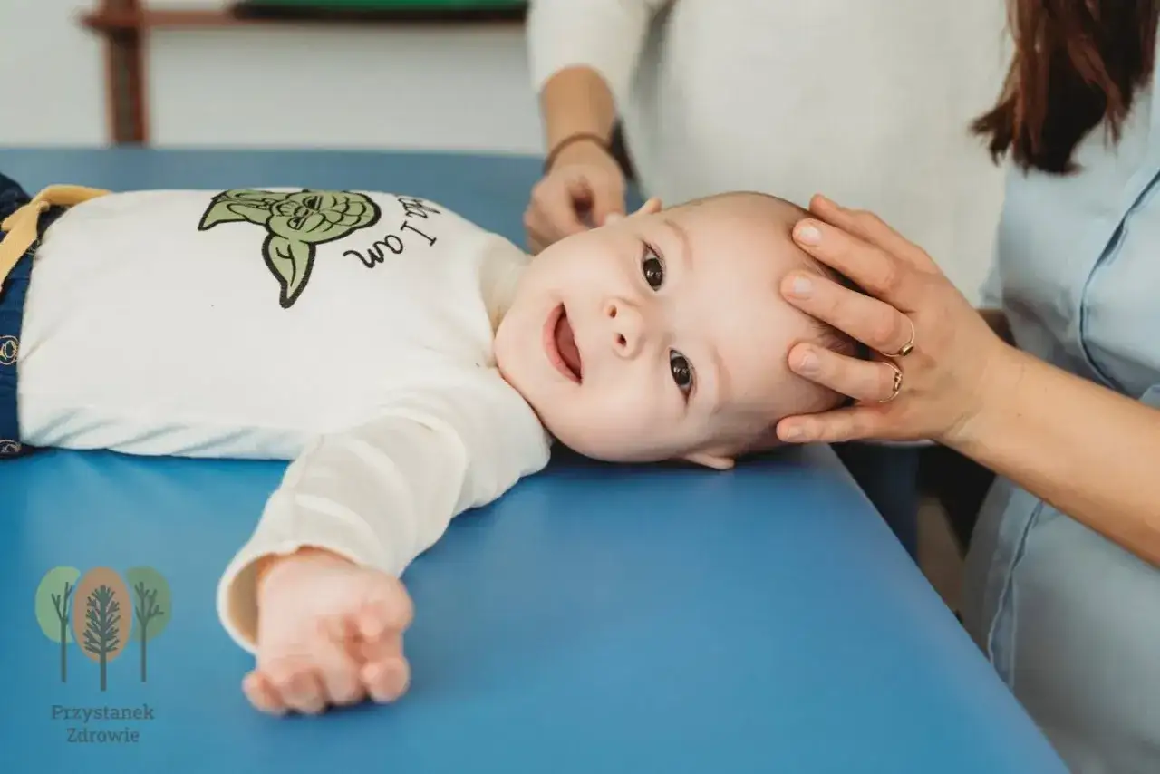 Uśmiechnięte niemowlę na niebieskim podłożu, ręce terapeuty delikatnie dotykają jego głowy. To wizyta u osteopaty dla dzieci.