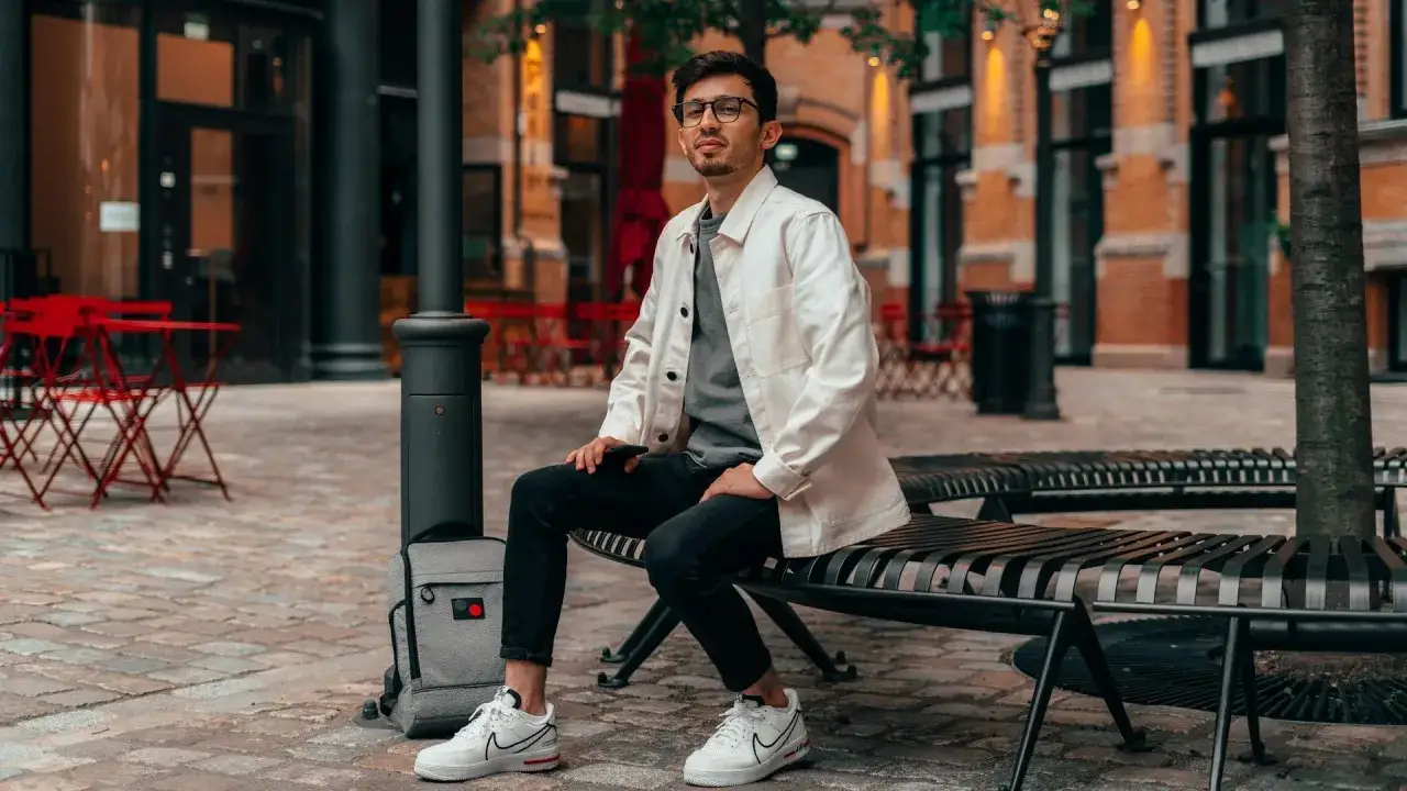 Mann in smart casual sommer outfit, sitzt auf einer Bank vor einem Geb&auml;ude mit roten Tischen.