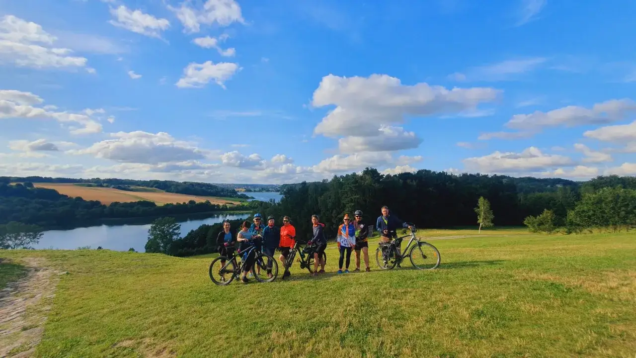 Grupa rowerzystów podziwia krajobraz, odkrywając najpiękniejsze trasy rowerowe na Kaszubach. Jezioro i pola w tle.