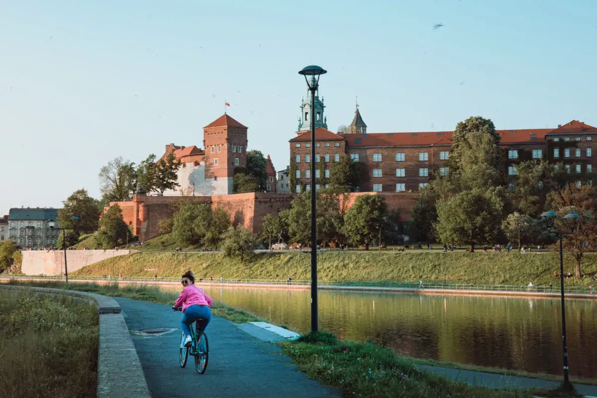 Kobieta na rowerze przemierza ścieżkę nad rzeką, w tle majestatyczny Wawel. Idealny dzień na wavelo.