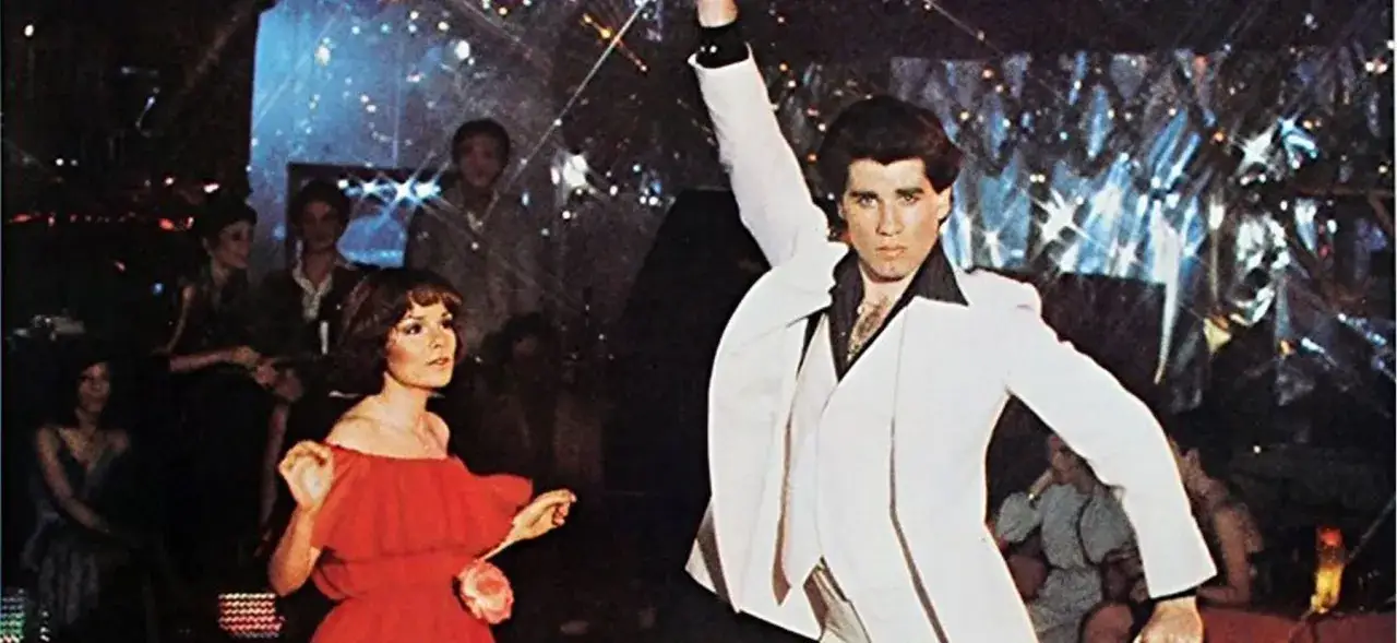 John Travolta w białym garniturze tańczy na parkiecie, a obok niego kobieta w czerwonej sukience. To kwintesencja gorączki sobotniej nocy.