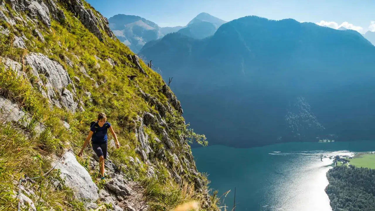 Wanderin auf einem steilen Pfad mit Seilsicherung, Blick auf den K&ouml;nigssee. Die K&ouml;nigssee Umrundung bietet atemberaubende Ausblicke auf Berge und See.
