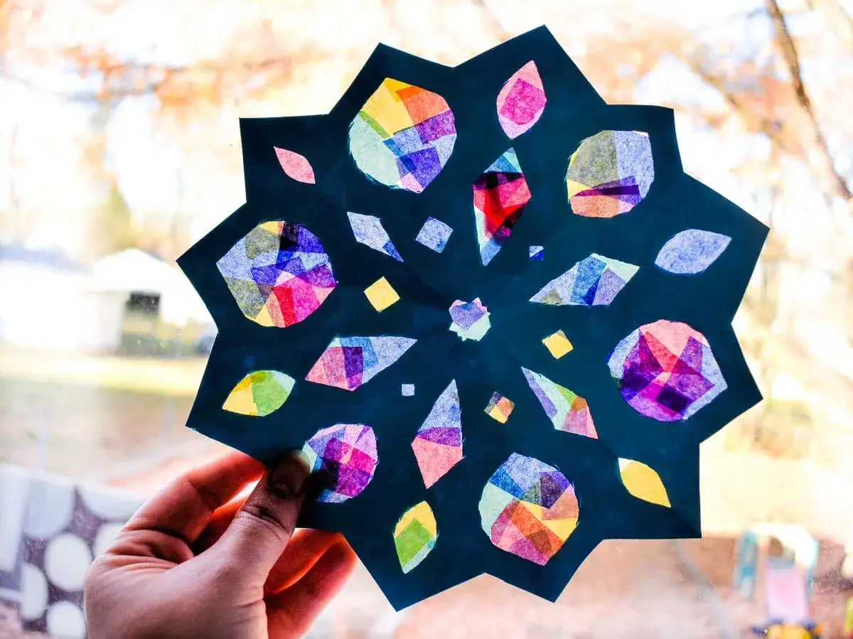 Bunte Schneeflocken basteln Kindergarten: Ein Stern aus Papier mit bunten Seidenpapierstücken, gehalten von einer Hand vor einem Fenster.