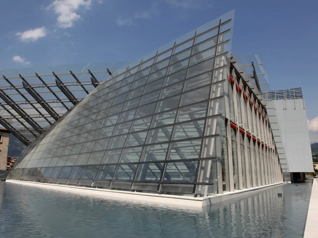 Il **Museo delle Scienze di Trento** si specchia in una vasca d'acqua, con la sua audace architettura in vetro e acciaio che cattura il cielo azzurro.