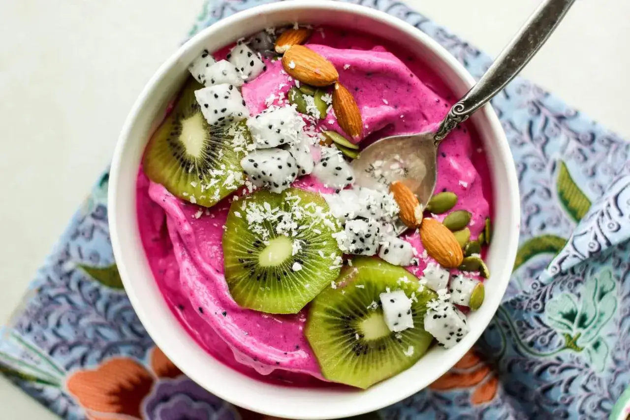 En rosa acai bowl med kiwi, drakfrukt, mandlar och pumpafrön. En sked vilar i skålen.