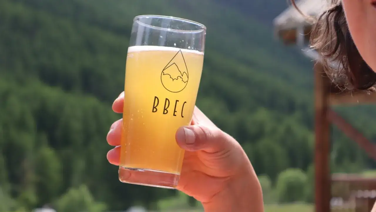 Verre de bière blonde pétillante, logo BBEC, avec un paysage de montagne verdoyant en arrière-plan. Parfait pour une bière printemps.