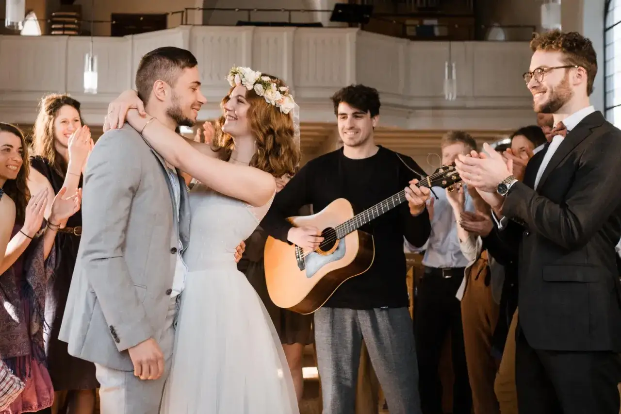 Glückliches Brautpaar umarmt sich, während ein Freund Gitarre spielt und Gäste applaudieren. Ein schöner Moment mit umgeschriebenen Liedern zur Hochzeit.