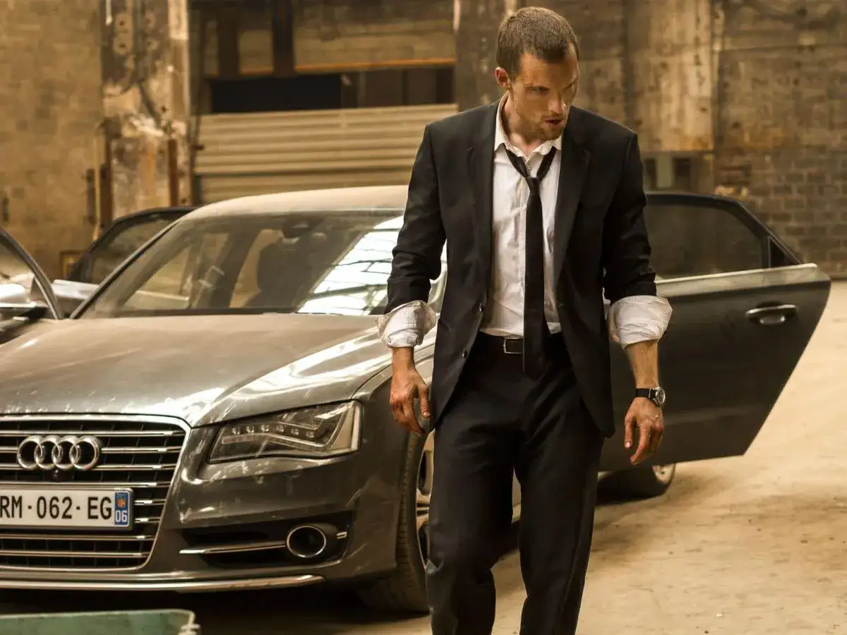 Ed Skrein, który grał Transportera, wychodzi z luksusowego Audi w garniturze.