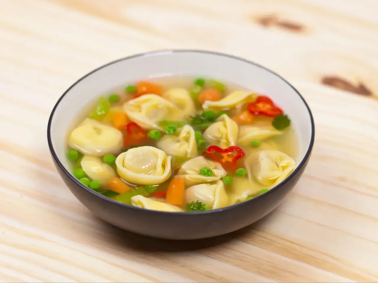 Miseczka gorącej zupy z tortellini na obiad, pełna warzyw: groszku, marchewki i papryczki chili.