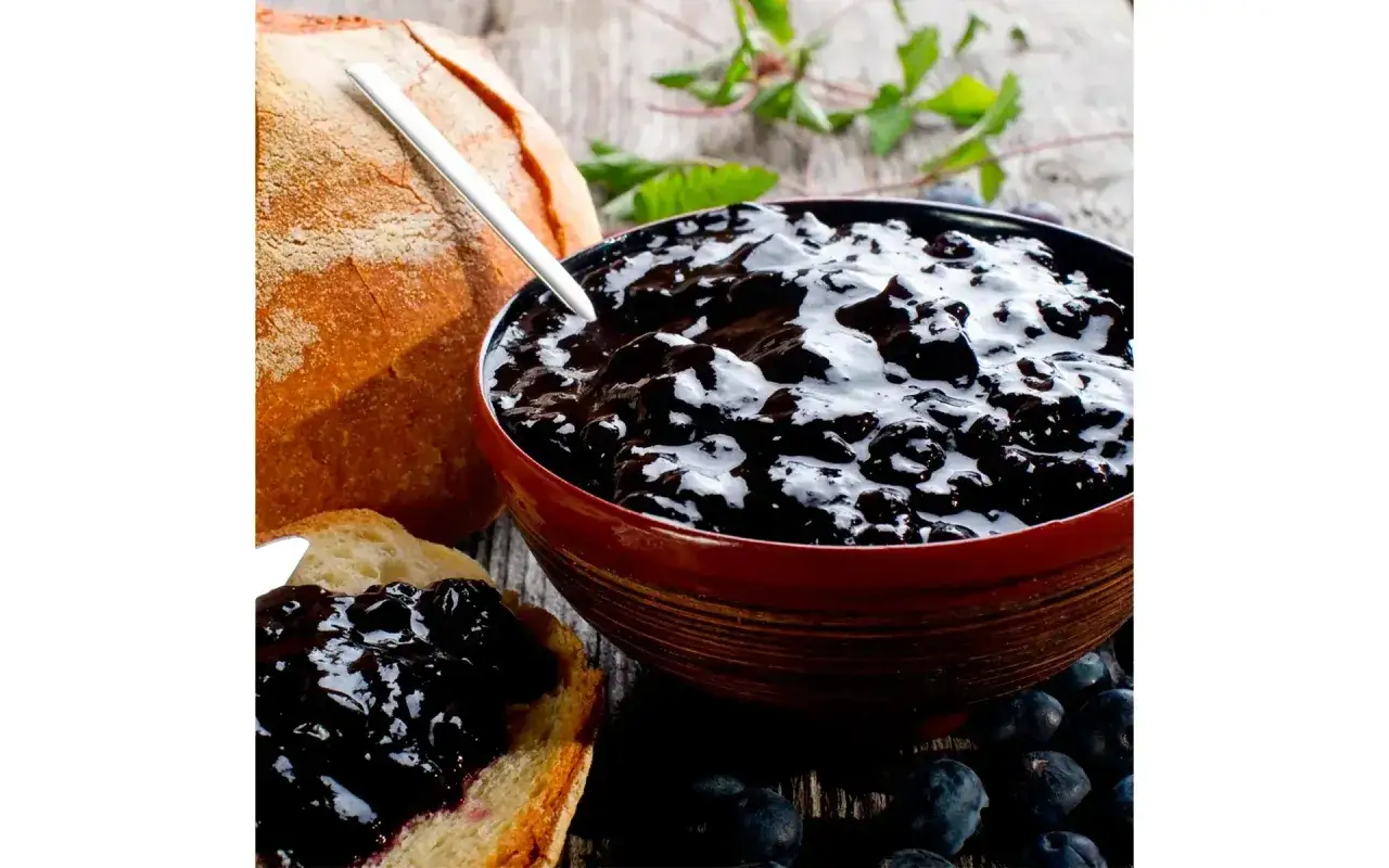 Una ciotola colma di marmellata di mirtilli, pane rustico e mirtilli freschi su un tavolo di legno.