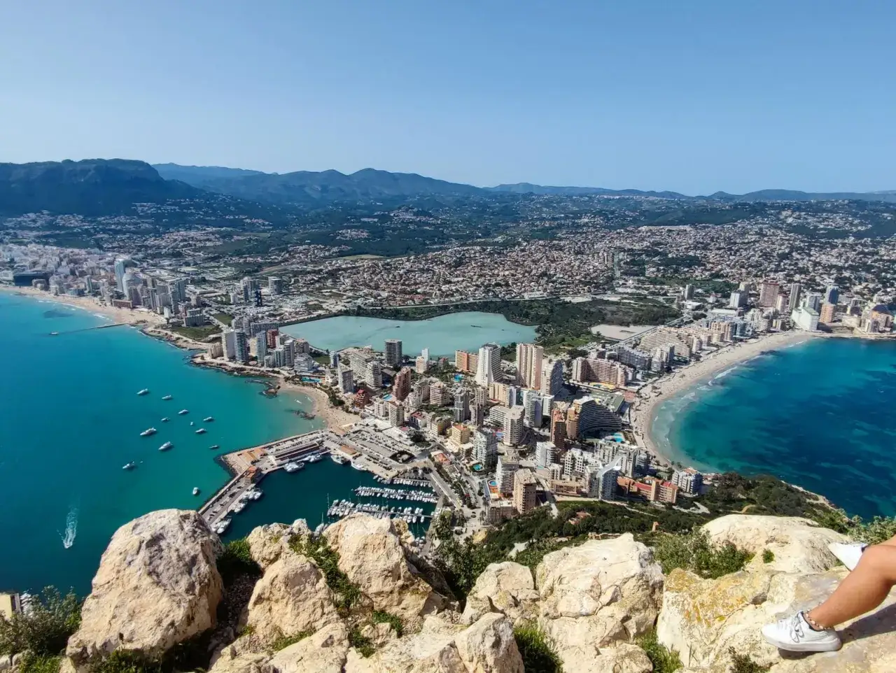 Vista aérea de la cala del penyal, con el mar turquesa, el puerto deportivo, la playa y la ciudad extendiéndose hasta las montañas.