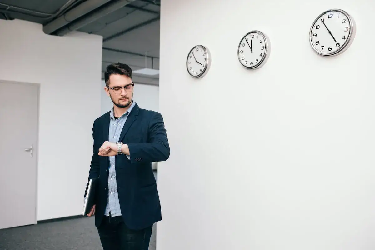 Mężczyzna w garniturze sprawdza zegarek, obok wiszą trzy zegary pokazujące różne strefy czasowe. Jego dyspozycyjność jest kluczowa w międzynarodowym biznesie.