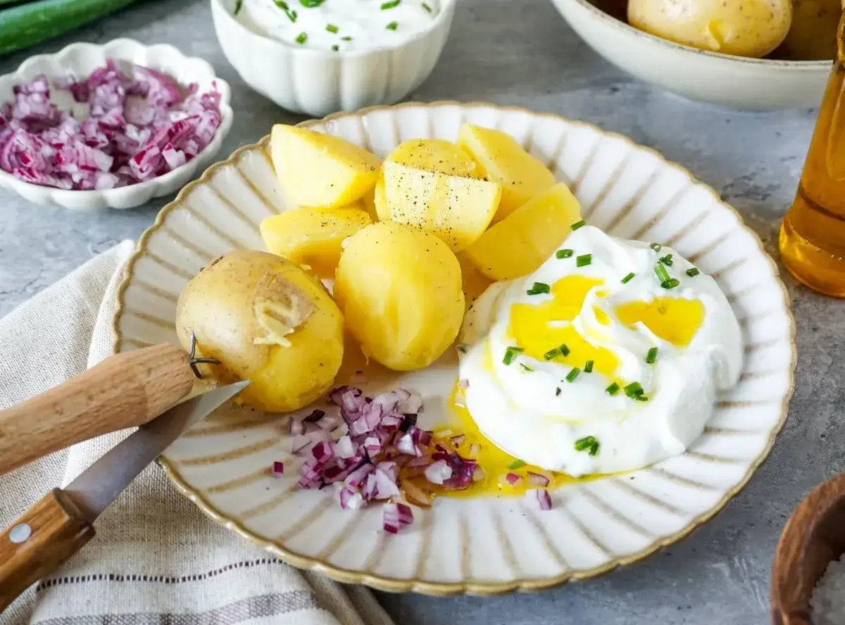 Salzkartoffeln mit Quark, garniert mit Schnittlauch und einem Schuss &Ouml;l. Dazu gibt es gehackte rote Zwiebeln und ein kleines Messer.