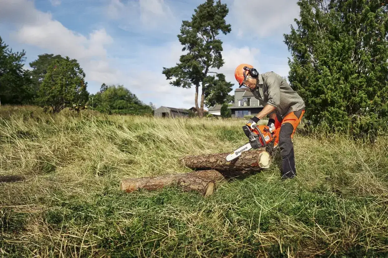 Mężczyzna w kasku i słuchawkach tnie piłą spalinową Husqvarna 445 leżące na trawie kłody. Dane techniczne tej piły sprawiają, że praca jest efektywna.