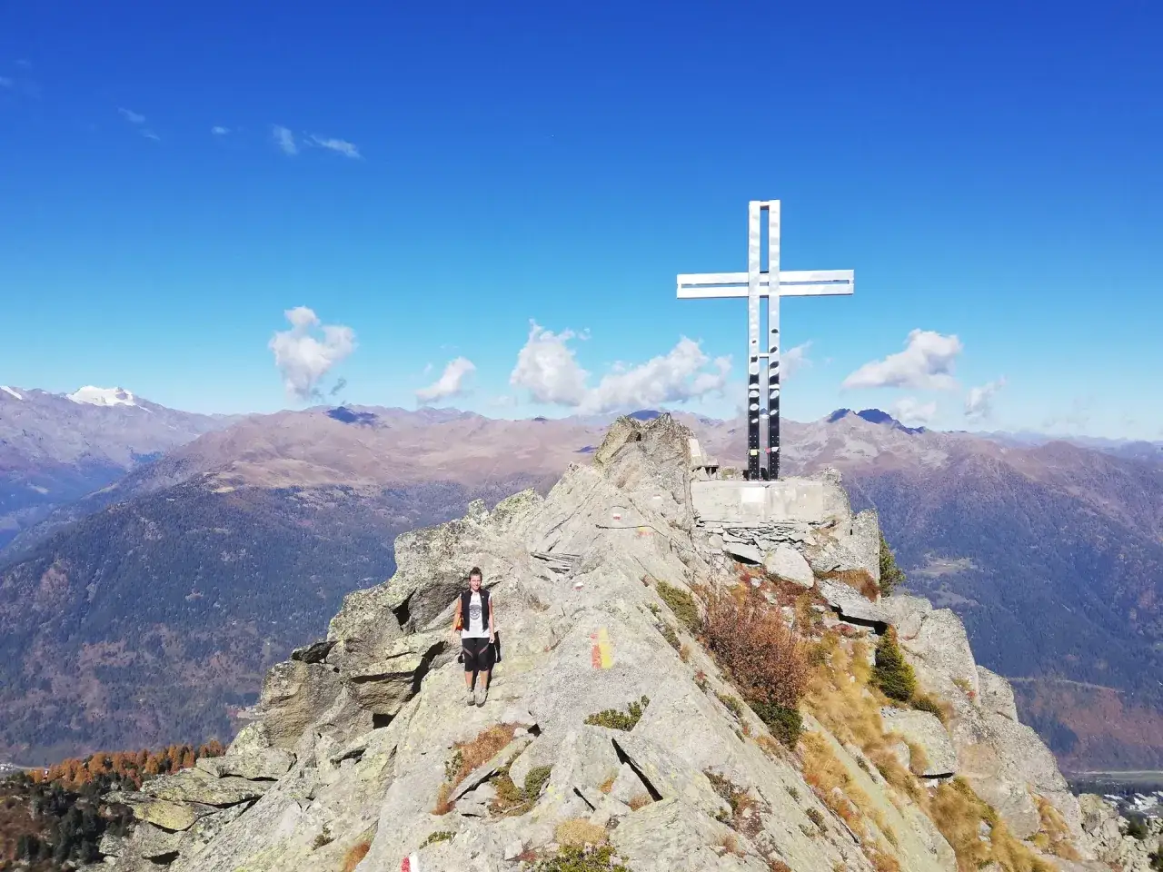 Escursionista raggiunge la vetta rocciosa dominata da una grande croce della pace, con un panorama alpino mozzafiato sotto un cielo azzurro.