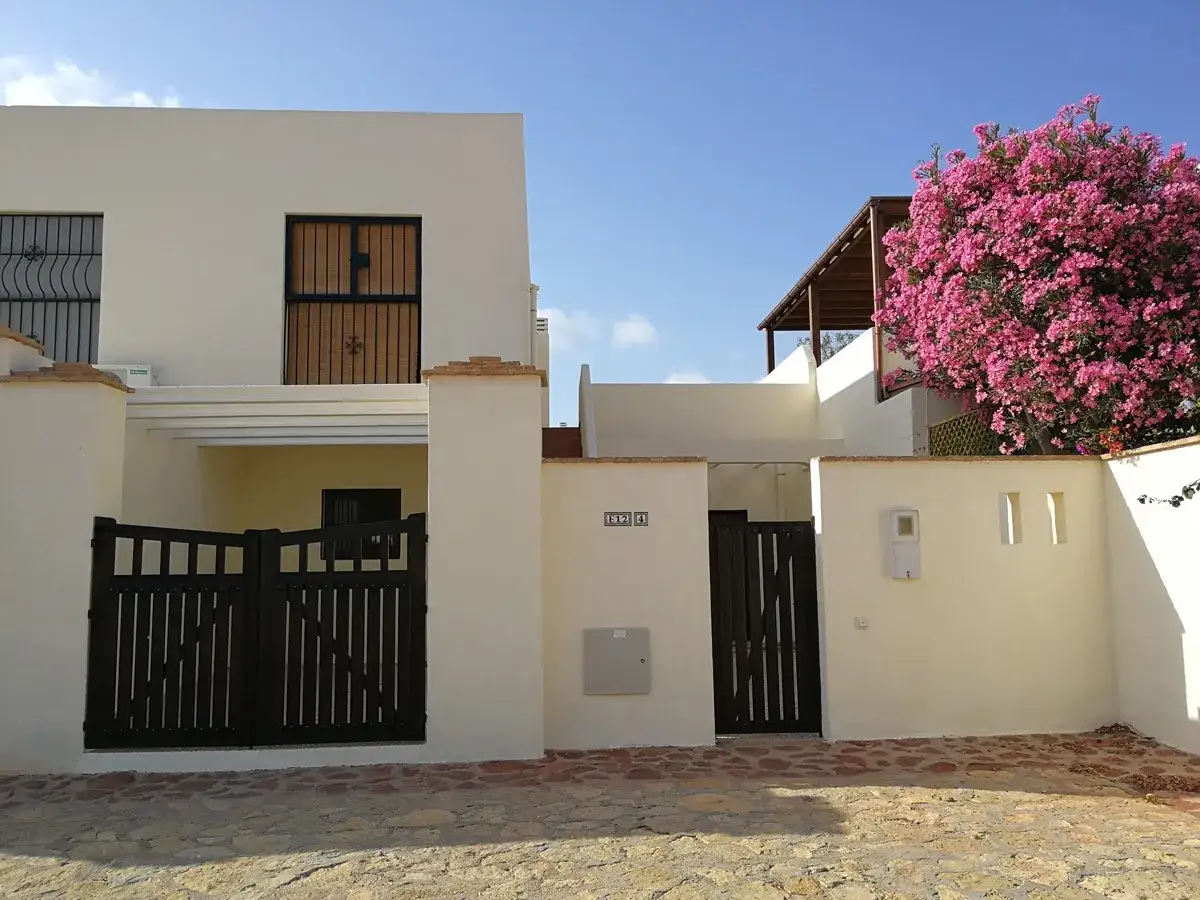 La casa en el sotillo San José, con puertas de madera oscura y un vibrante árbol de flores rosas, bajo un cielo azul.
