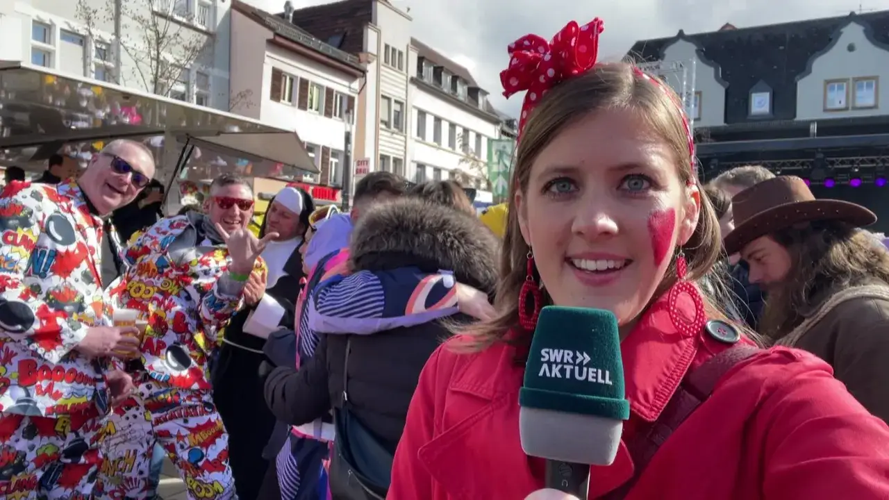 Am **Donnerstag Karneval** feiern Menschen in bunten Kostümen, darunter Comic-Muster und Nonnen. Eine Reporterin hält ein Mikrofon.