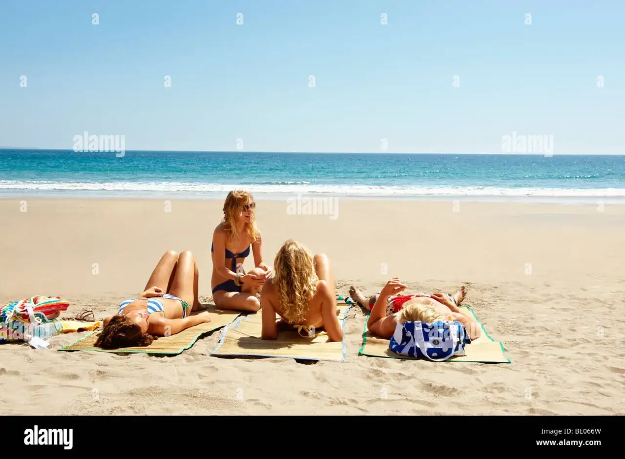 Quatre amies profitent du soleil sur la plage, allong&eacute;es sur des nattes pour se faire dorer la pilule. L'oc&eacute;an bleu s'&eacute;tend &agrave; l'horizon.