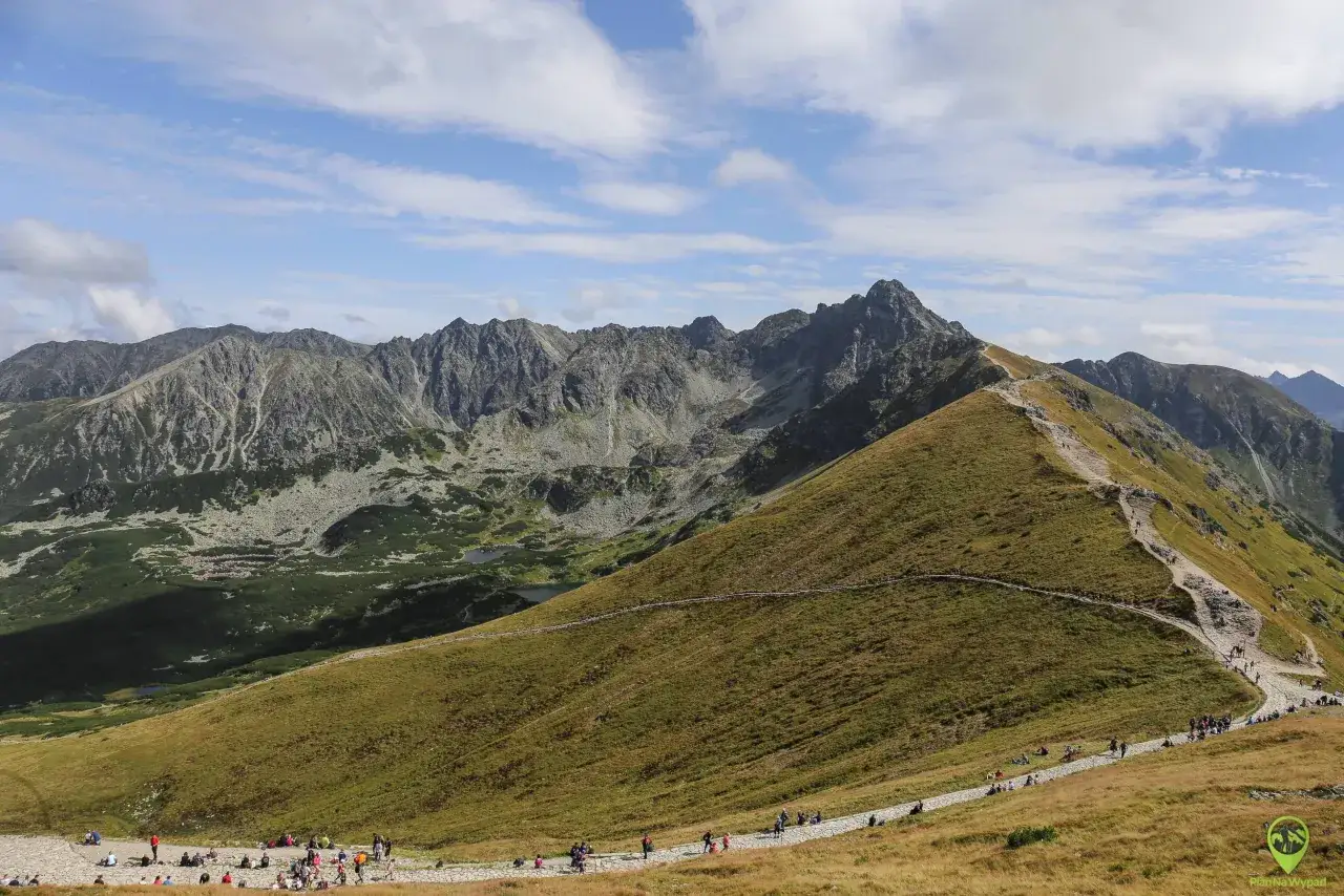 Widok na Tatry z kamienistym szlakiem, kt&oacute;rym podążają turyści. To łatwe zejście z Kasprowego Wierchu, idealne na spacer.