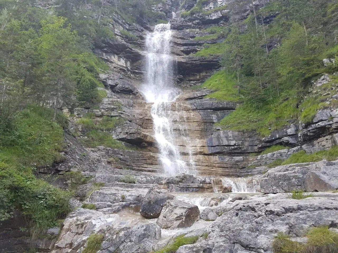 Ein tosender eibsee wasserfall stürzt über Felsen in ein steiniges Bachbett. Grüne Vegetation umrahmt die Szenerie.